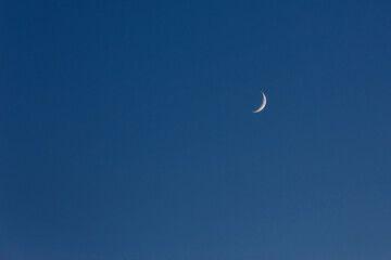 A delicate thin crescent moon hangs in a clear deep blue evening sky, a natural celestial sight. © tai