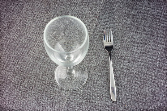 A clear empty stemmed glass goblet and a metal fork rest on a gray woven textured surface. Simple tableware arrangement.