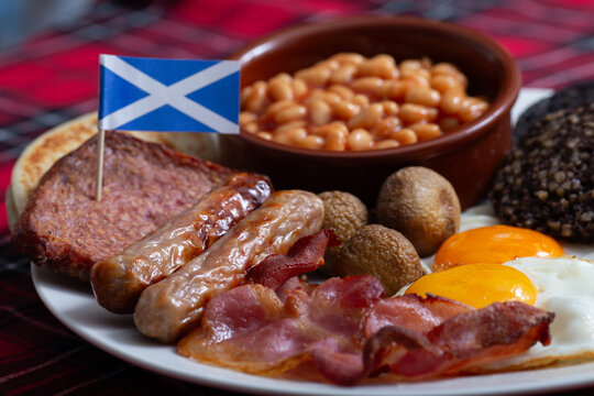 Traditional Full Scottish Breakfast plate featuring Lorne sausage, haggis, black pudding, grilled tomatoes, fried eggs, mushrooms, crispy bacon, sausages, baked beans, and tattie scones with a mini Sc