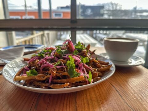 A large white plate filled with shoestring French fries, deep-fried and crispy, covered with cheese sauce, pickled onions, and cilantro. There&rsquo;s a white porcelain cup and saucer on the table.