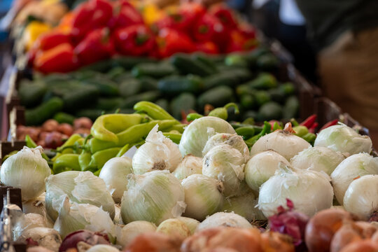 A farmers' market with bins of various colorful fresh vegetables. There are white and red onions, green, banana, red and yellow peppers, shallots, and avocados.  The shopping bins are full.