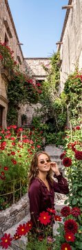 Young woman standing in a narrow stone alleyway filled with blooming red flowers