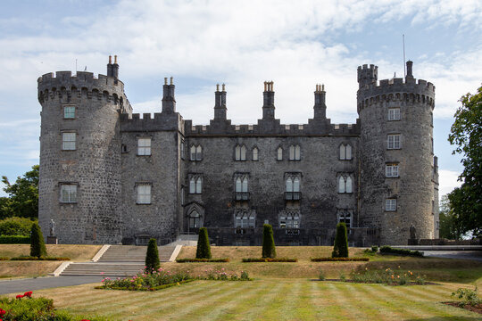 Symmetric front view of Kilkenny Castle with stone towers and formal rose garden terrace under cloudy sky in Kilkenny, Ireland