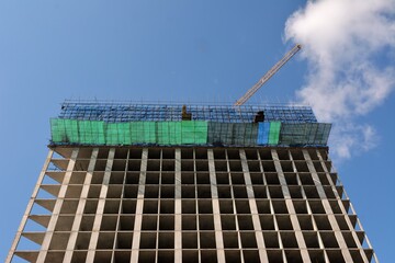 A view from below of the reinforced concrete frame of a high-rise building under construction, with fencing mesh and the boom of a construction crane against a cloudy sky. © Алексей Желтухин