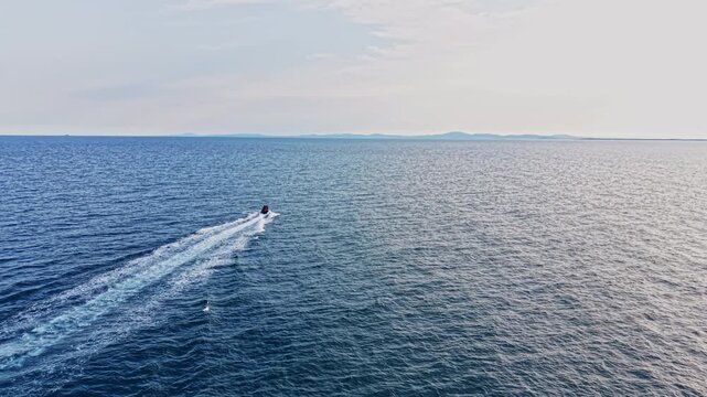 A boat moves swiftly over the blue water. The ocean spreads wide with gentle waves. The sky is bright, and land appears far away on the horizon.
