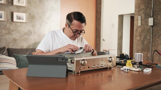 Mature Asian man disassembling audio equipment and self-learning, searching for manual guide to fix circuit board in tablet, home appliance DIY, electronics component hobby, handyman skill workshop.