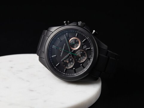Elegant black chronograph wristwatch resting on a white marble surface against a dark background.
