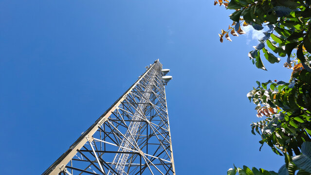 Low angle shot of a tall steel telecommunication tower reaching into a vibrant blue sky framed by green tree leaves on the side.