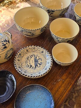 Handmade cream ceramic bowls and plates with hand-painted blue crab and floral motifs sit on a rustic wooden table, including a lidded tureen and smaller serving pieces. The collection has a warm, art