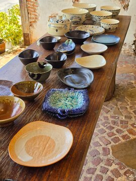Handmade cream ceramic bowls and plates with hand-painted blue crab and floral motifs sit on a rustic wooden table, including a lidded tureen and smaller serving pieces. The collection has a warm, art