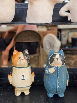 Handmade wooden cat figurines in playful painted outfits stand in a row on a market shelf, each carved with chunky, characterful shapes.
