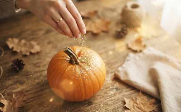 Woman decorating a small autumn pumpkin with golden glitter on a rustic wooden table surrounded by dry leaves for seasonal craft and DIY projects