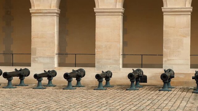 A row of historic bronze military cannons neatly lined up on cobblestones beneath classical stone arches inside the courtyard of Les Invalides museum in Paris, France during daytime.

