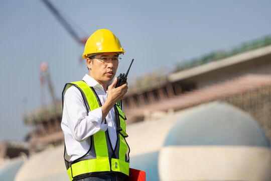 Asian construction engineer in hard hat and safety vest talking walkie talkie on site, Site supervisor using a radio communication device at industrial infrastructure project