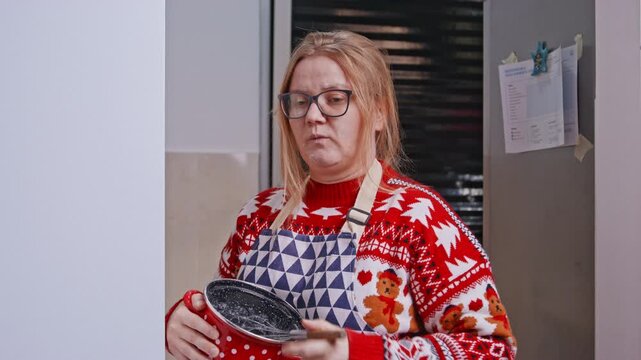 An adult woman wearing a festive sweater and apron holds a mixing pot and whisk in a kitchen, then pauses to check a recipe on her smartphone.