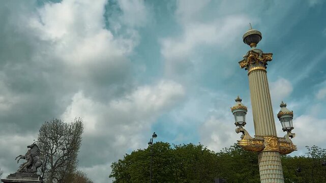 Tourists and pedestrians walk past an ornate gilded lamppost column and historic equestrian sculpture on Place de la Concorde beside Tuileries Garden in central Paris, France, under clouds.

