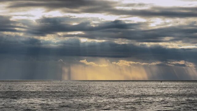 Golden sunbeams pierce heavy storm clouds above a rippling ocean, with distant rain showers visible on the horizon at sea.