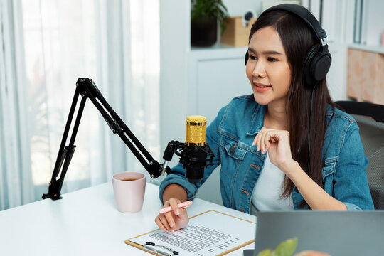 Host channel Asian influencer talking in broadcast streaming online wearing headset on social media live with script note, greeting listeners in life coaching or marketing at modern studio. Stratagem.
