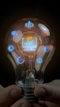 Close-up vertical shot of hands holding lightbulb with glowing KPI monitor and growth metric icons. Corporate business performance evaluation, strategic goal benchmarking, and management concept.