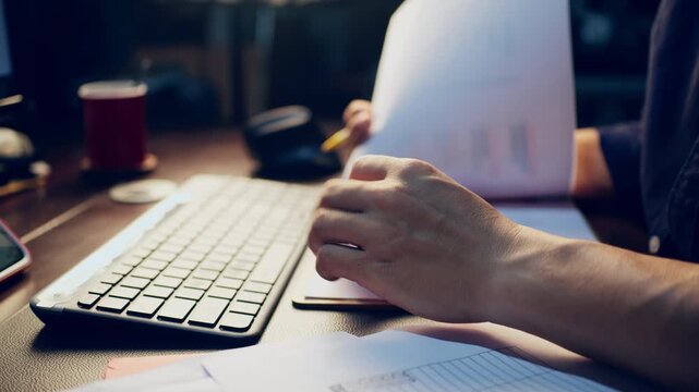 Close-up of a businessman checking financial data on clipboard documents and typing on a computer keyboard at his desk.