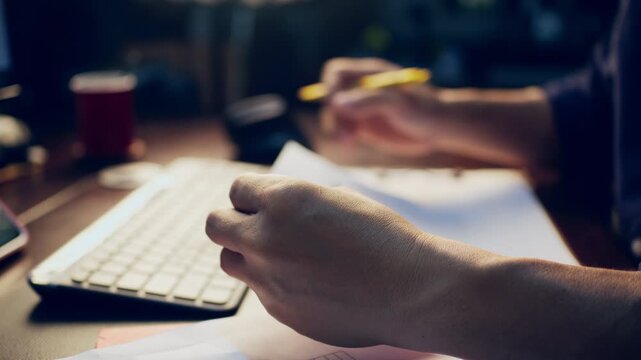 Close-up of a businessman checking financial data on clipboard documents and typing on a computer keyboard at his desk.