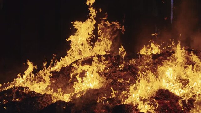 Medium-close static shot capturing tall, vigorous tongues of yellow and orange fire dancing across an expansive bed of dark smoldering ash and burning joss paper remnants.