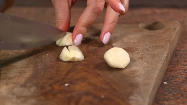 Close up of hands chopping peeled garlic with a kitchen knife on a rustic wooden cutting board while preparing ingredients for a homemade meal.