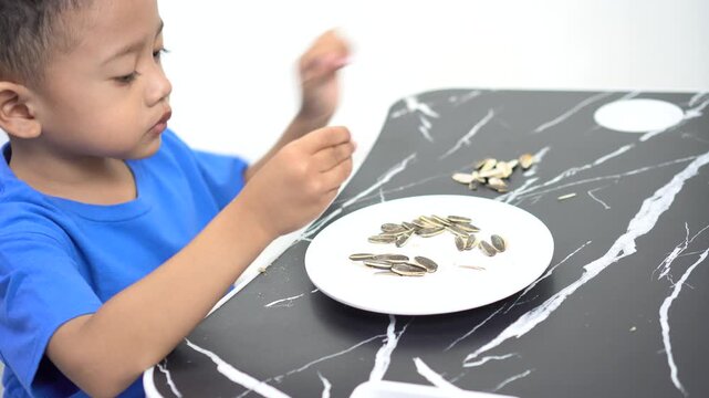 A handsome Asian boy is intently shelling and eating sunflower seeds, healthy snack for children, isolated on a white background.