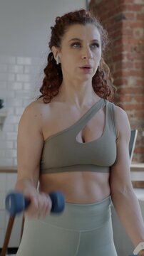 Strong young lady in sports outfit training with dumbbells indoors in apartment focused on bodybuilding. Sportswoman and house concept.
