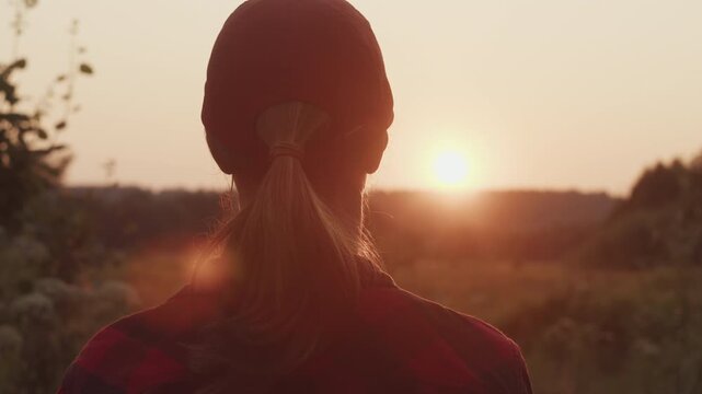 Young woman in a red cap watches the sunset over a rural landscape, viewed from behind.