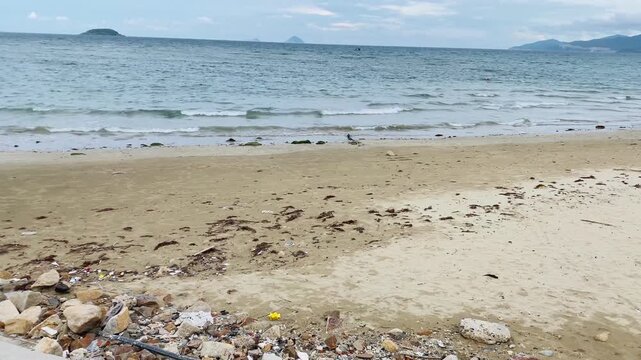 Environmental pollution on a sandy sea beach with plastic waste, construction debris, and microplastics washed ashore near the water.