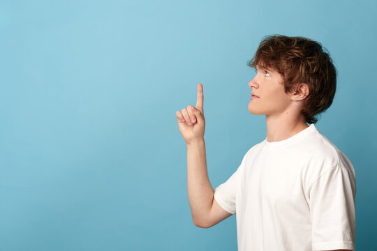 Thoughtful young man in white tshirt raising index finger and looking up in profile on a blue background. Concept of having an idea, inspiration, and creative thinking.