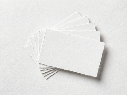 A stack of small, rectangular white paper cards with deckled edges fanned out on a textured white background, viewed from above.