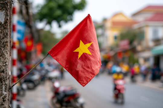 Vietnamese flag waves gently in street breeze. Colorful buildings blur softly in background. Motorbikes move through busy urban scene. Calm, vibrant daily life captured in vivid detail