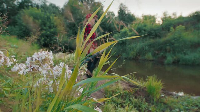 Young adult woman walks with fly fishing rod on the lush green coast to the small river