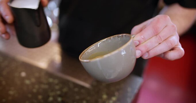Latte art. Pouring foamed milk. Barista hand adding frothing creamy component from pitcher to beverage garnished coffee in ceramic cup.