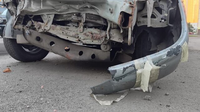 Severely damaged front bumper and detached body parts of a car after a serious road accident. Close-up view of deformed metal structures and broken elements on the asphalt road.