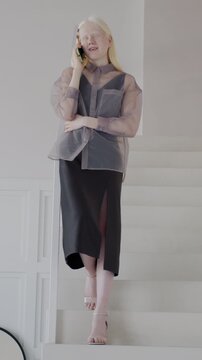 Elegant albino woman speaking on mobile phone standing on stairs in luxury house. Communication and stylish apartment interior concept.