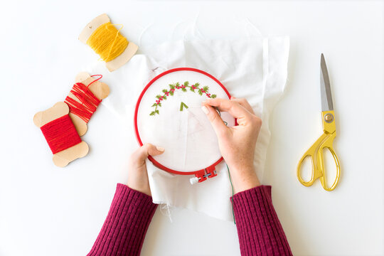 Hands embroidering with red thread, surrounded by sewing tools.