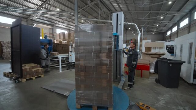 Industrial warehouse packaging line with tall wrapped pallet of stacked cardboard sheets on blue turntable beside vertical wrapping machine inside spacious warehouse