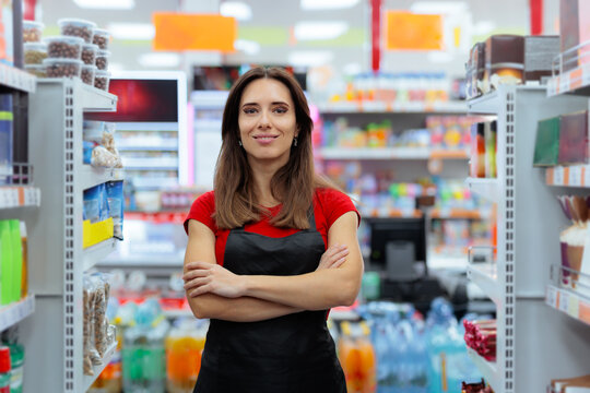 Confident Female Supermarket Worker Standing With Arms Crossed. Smiling female grocery store employee wears a black apron in a supermarket aisle.
