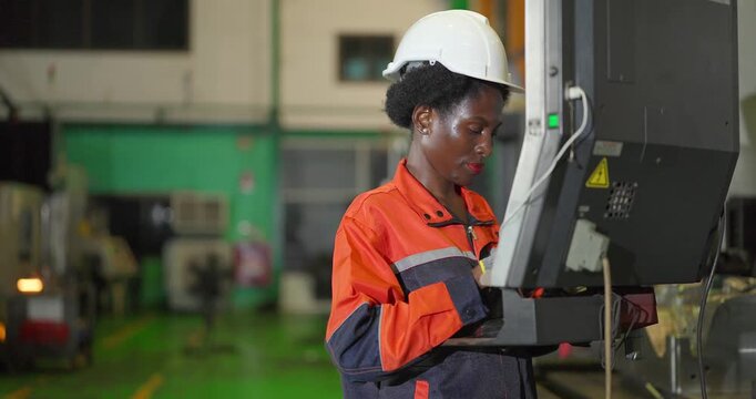 A Black female machine operator in orange work coveralls and a white hard hat enters settings on the keyboard of a CNC machine control panel on the floor of a metal parts factory.
