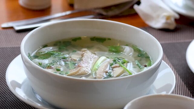 Bowl of hot vietnamese pho soup with chicken and herbs steaming on table in restaurant.