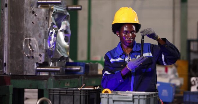Female factory worker carefully examines a long bolt before assembly beside an industrial mold. Wearing a yellow hardhat, gloves, and blue workwear.
