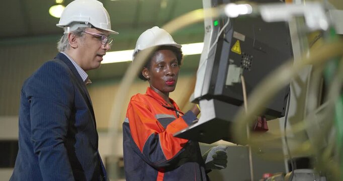 A senior manager in a suit and hard hat stands with a Black female machine operator at a CNC machine control panel, discussing settings and checking production on the factory floor.
