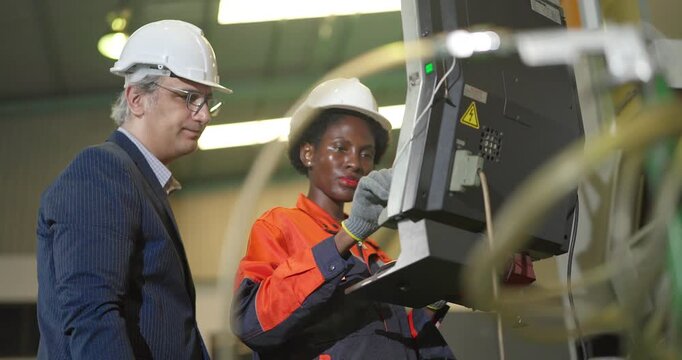 A senior manager in a suit and hard hat stands with a Black female machine operator at a CNC machine control panel, discussing settings and checking production on the factory floor.
