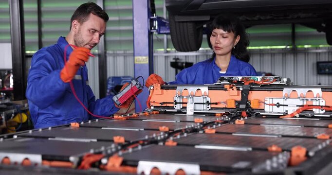 Two automotive technicians use diagnostic equipment to inspect an electric vehicle battery pack in a repair workshop. Wearing blue workwear and orange protective gloves.