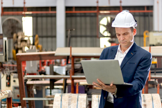 Portrait of happy senior business owner in suit working with laptop in manufacturing factory. Manager with white helmet using computer during working and inspecting machines at industrial site.