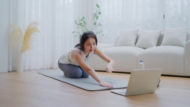 Young Asian woman stretching yoga workout on exercise mat while online training class with computer laptop in living room
