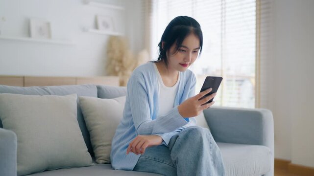Smiling pretty young Asian woman sitting on cozy couch, using funny mobile apps in living room. Woman at home, Doing online shopping, Messaging friends, Posting on social media
